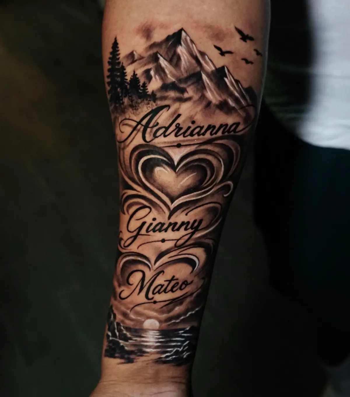 Black and grey forearm tattoo combining mountains, layered hearts, script lettering, and a waterside landscape