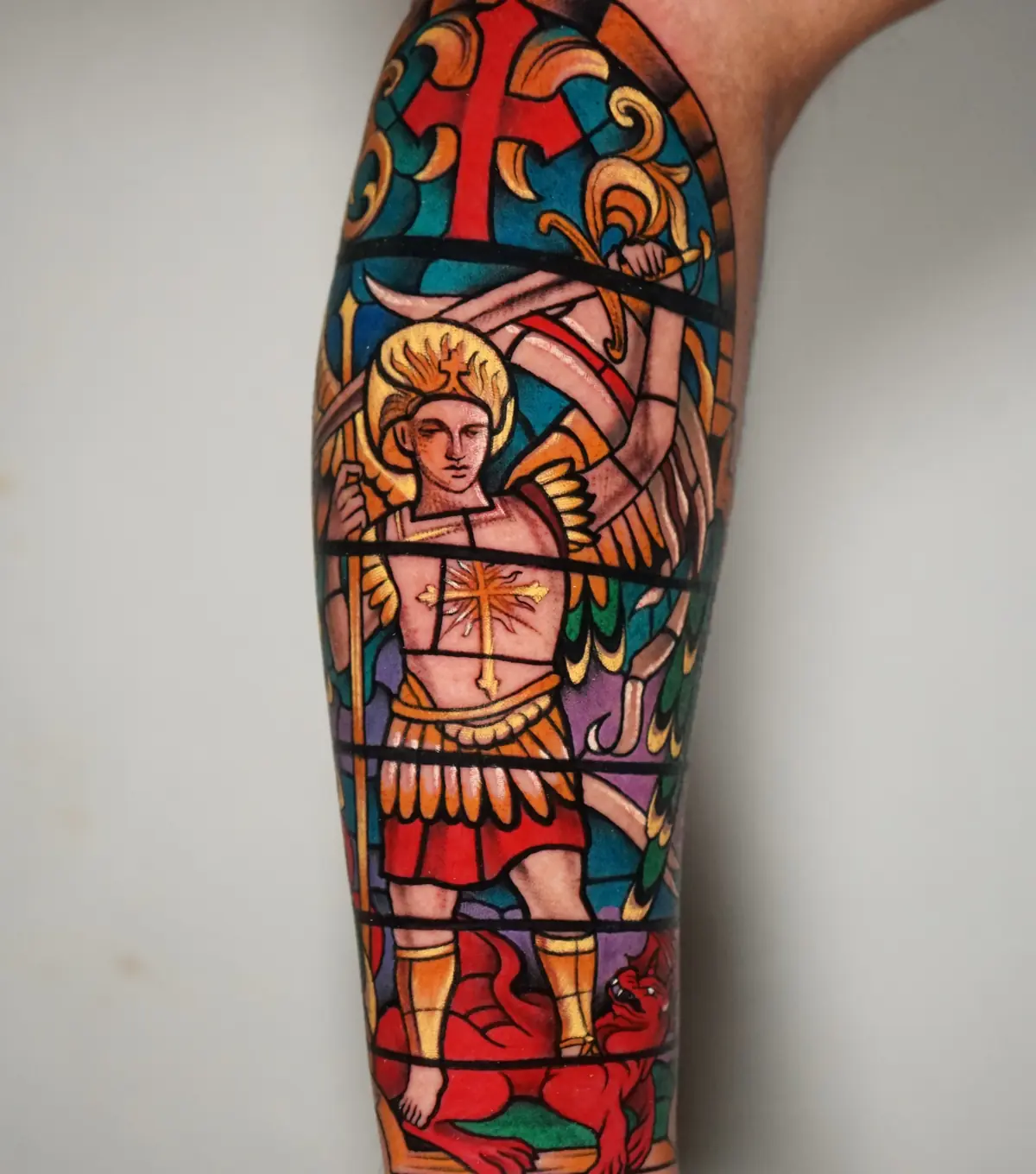Traditional stained-glass style tattoo of Saint Michael with bold red, blue, gold, and green sections